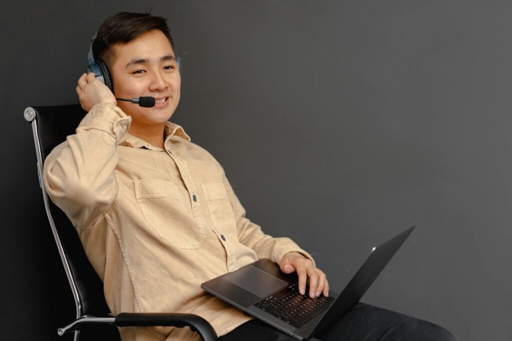 An Asian man seated on a chair, using a laptop while wearing a headset, focused on his work.